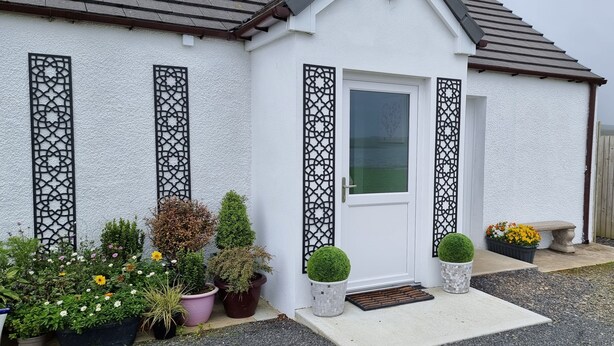 Une maison blanche avec une porte d’entrée, des panneaux noirs décoratifs, des plantes en pot et une clôture en bois.