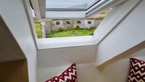 Une vue depuis une fenêtre à cadre blanc, donnant sur un jardin avec une clôture en bois, un petit arbre et un parterre de fleurs circulaire.