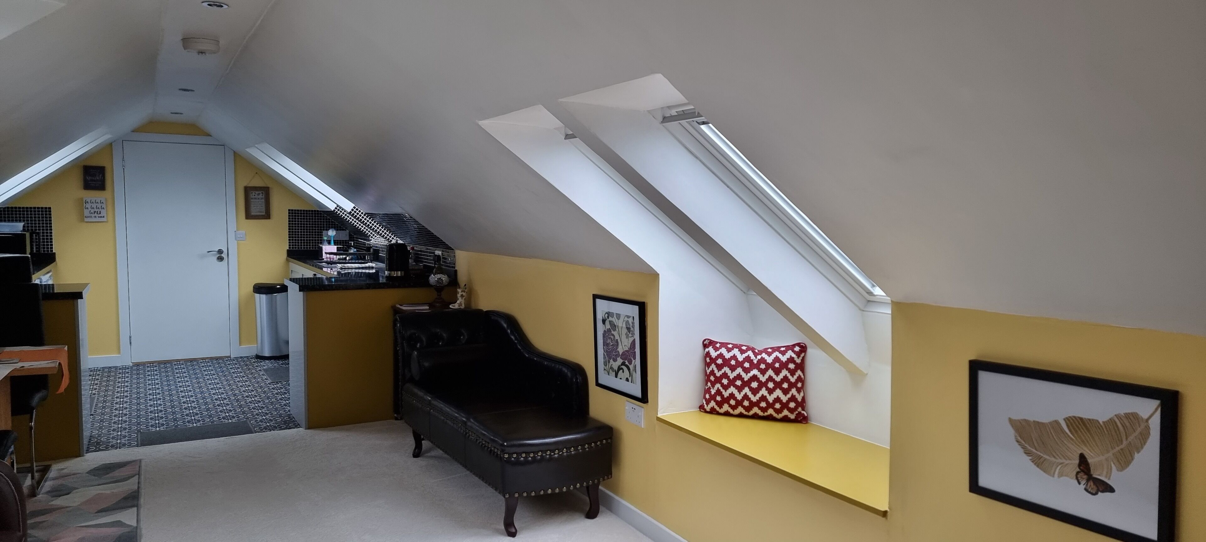 Una habitación con techo inclinado, una butaca de cuero negro, una pared amarilla y un cuadro con una mariposa.