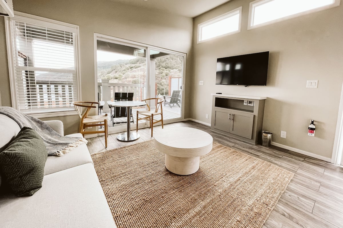 A modern living room with a sofa, a round coffee table, and a TV mounted on the wall. There is a balcony with a view of the outdoors.