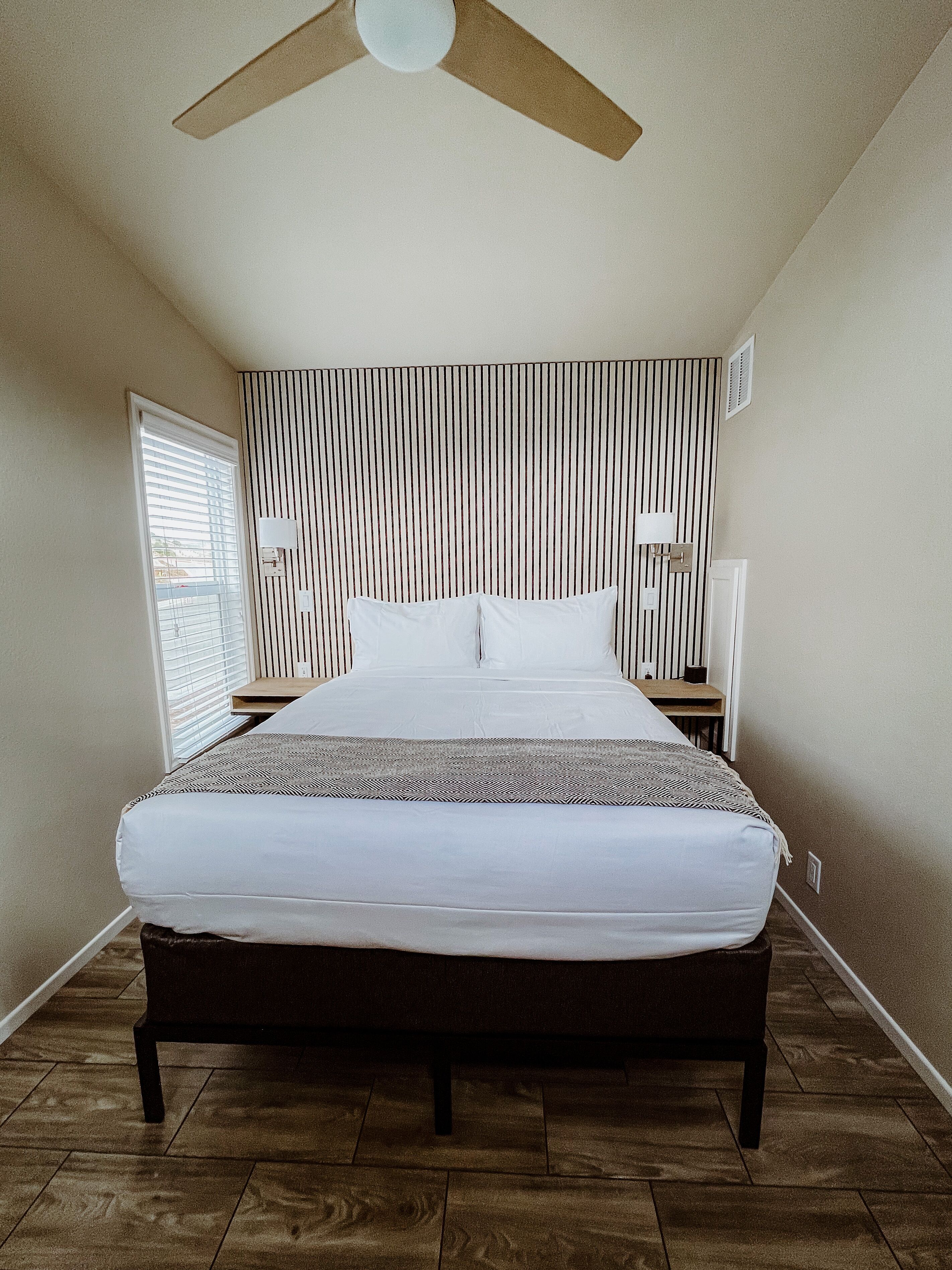 A bedroom with a bed, a ceiling fan, a window with blinds, and a wall with vertical lines.