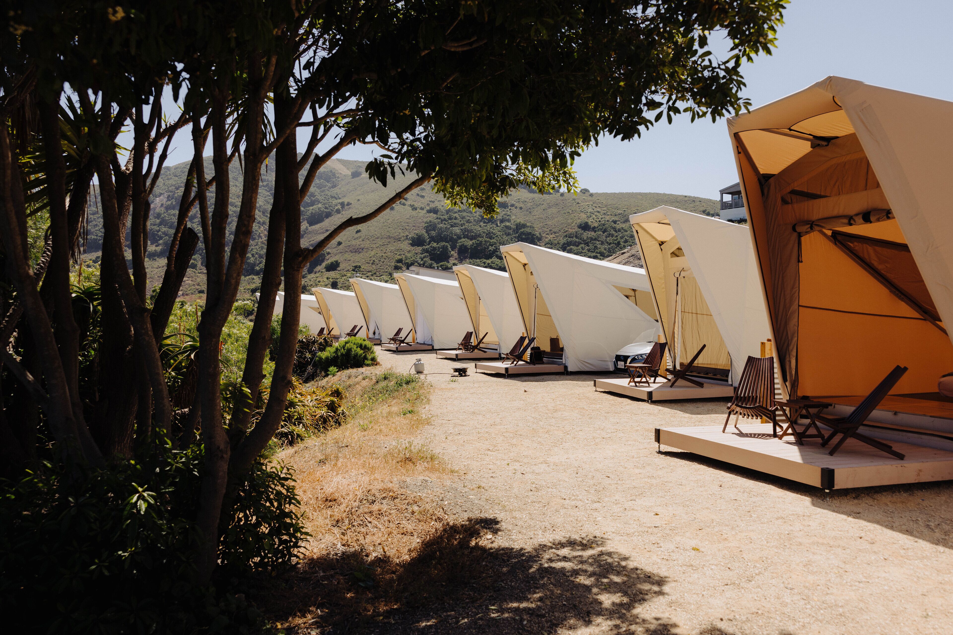 A row of tents with attached chairs and tables, set in a natural landscape with trees and hills in the background.