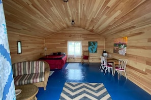 A cozy cabin with a wooden interior, a bed, a sofa, a dining table with chairs, and artwork on the walls.