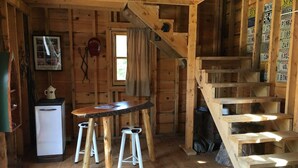 Interno di una baita rustica con un tavolo in legno, sgabelli, un piccolo frigorifero, una finestra con tende e una scala in legno.