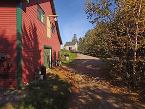 A red wooden house with a green door and windows, a driveway leading to a white building, and trees with autumn leaves.