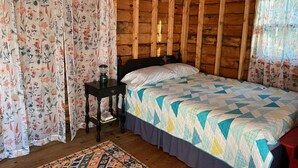 A wooden cabin bedroom with a bed, a nightstand, a lamp, and floral curtains.