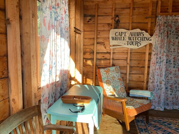 L’intérieur d’un chalet chaleureux, avec un mur en bois, une enseigne indiquant « Excursions d’observation des baleines du capitaine Achab », un panier en osier, une table bleue et un fauteuil à motifs.