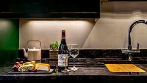 Una encimera de cocina con una botella de vino, dos copas, una tabla de quesos, una tetera y un lavabo.