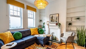 Una sala de estar moderna con un sofá modular, una mesita auxiliar, una lámpara de pie y una silla. Hay ventanas con persianas amarillas, una chimenea y estanterías con plantas.