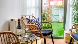 Una mesa de vidrio con sillas de mimbre y cojines, una botella de vino y vistas a un jardín verde a través de un ventanal.