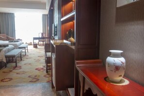 A hotel room with a bed, a desk, a chair, a small table with a vase, and a patterned rug.