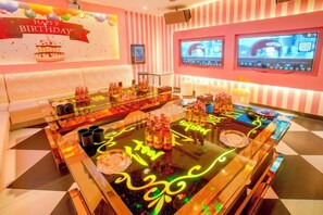 A bar area with a neon-lit table, cushioned seating, and a birthday-themed wall decoration.