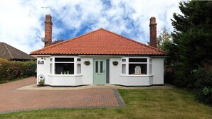 Une maison à deux étages, avec un toit recouvert de tuiles rouges, des murs extérieurs blancs et une porte d’entrée verte. Elle possède deux cheminées, un petit porche agrémenté de plantes en pot et un chemin de briques menant à l’entrée.