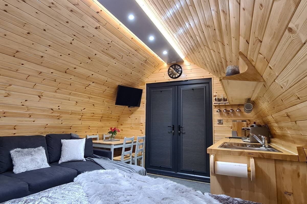 L’intérieur chaleureux d’un chalet avec un plafond en bois, une porte noire, un canapé avec des coussins, une petite table avec des chaises et un coin cuisine.