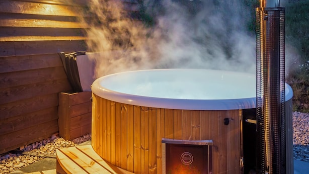 A hot tub with a wooden exterior and a chimney emitting steam, surrounded by a wooden deck and a stone pathway. - Loch Ewe Luxury Pods (Achnasheen)