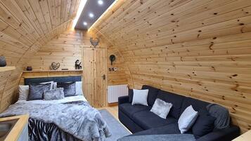 A cozy cabin interior with a wooden sloped ceiling, a bed with a fluffy blanket, a black sofa with pillows, and a small shelf with decorative items.