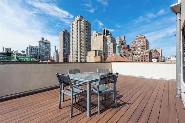 A rooftop terrace with a glass table and chairs, overlooking a city skyline with high-rise buildings.