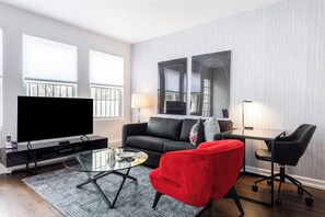 A modern living room with a large flat-screen TV, a black leather sofa, a red armchair, a glass coffee table, and a desk with a lamp.