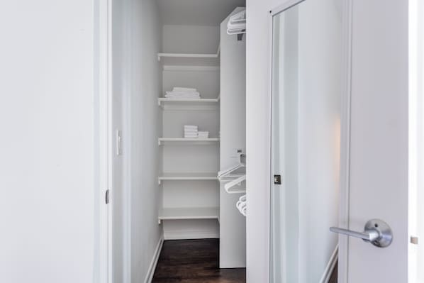 A narrow hallway with white walls and a wooden floor leading to a closet with white shelves and hanging storage.