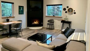 A modern living room with a fireplace, a sofa, a dining area, and large windows with a forest view. - The Treehouse Suite with a roof top jacuzzi hot tub. Private and Quiet. (Bonnington Falls)