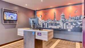 A hotel reception desk with a monitor displaying a map, a Dell computer, and a mural of a coastal cityscape. - Premier Inn Torquay Harbour (Torquay)