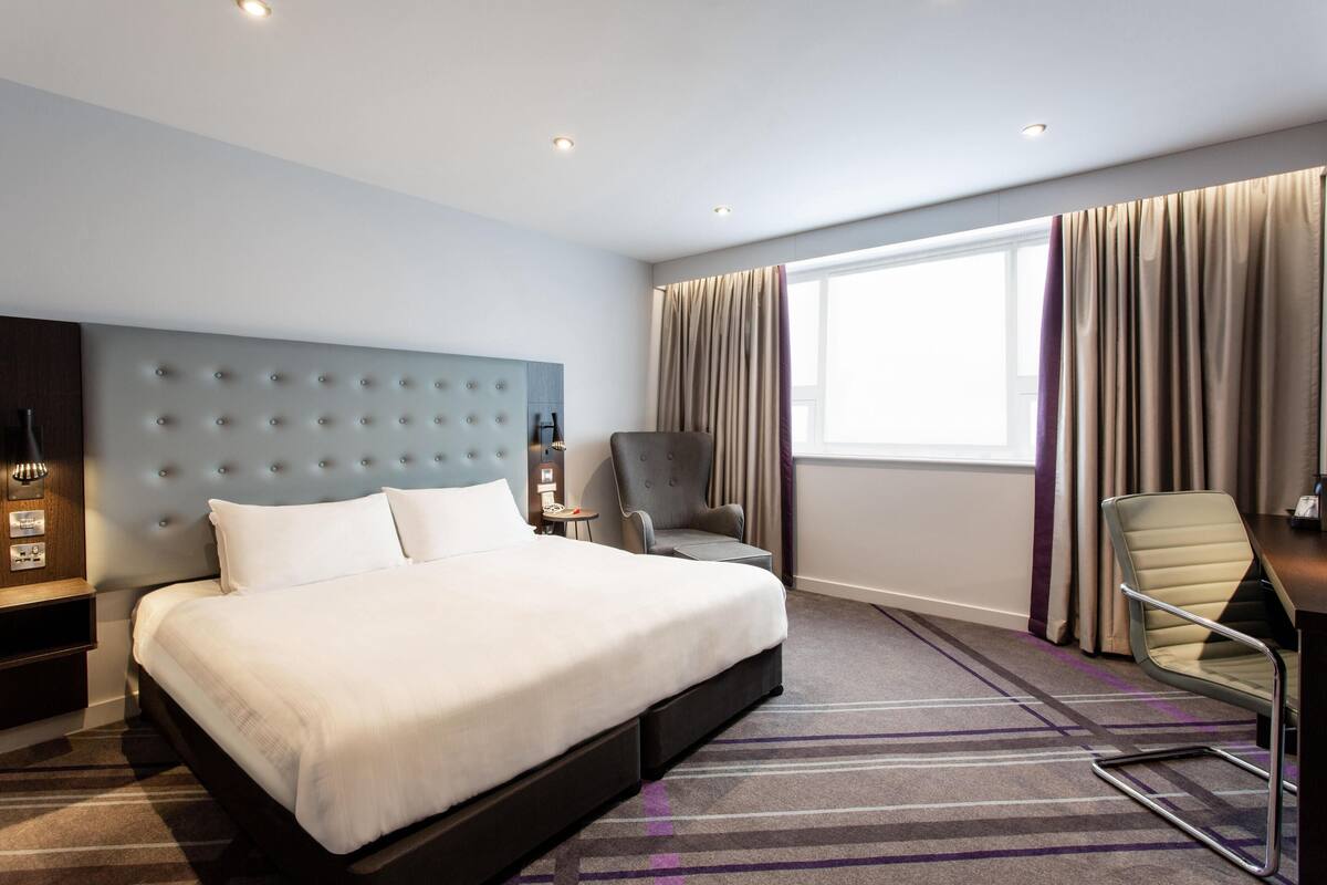 A hotel room with a large bed, a desk, a chair, a nightstand, and a window with curtains.