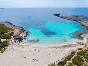 A coastal beach with clear turquoise waters, rocky outcrops, and a few boats.