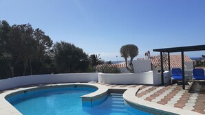 En pool med en böjd kant, ett kaklat poolområde och en överbyggd sittplats med blå stolar.