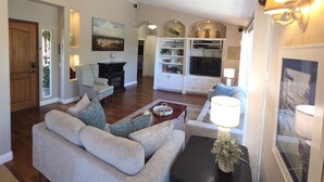 A spacious living room with a sofa, a coffee table, and a TV. There is a fireplace, a bookshelf, and a dining area visible in the background. - Serene Guest House on Avocado Grove and Vineyard (Fallbrook)