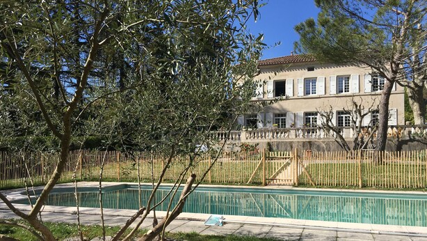 A swimming pool with a wooden deck, surrounded by a bamboo fence, and a large building with multiple windows and balconies in the background. - Self-contained apartment in a magnificent estate (Romans-sur-Isère)