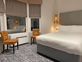 Een hotelkamer met een bed, twee stoelen, een raam met zonwering, een radiator en een wandlamp.