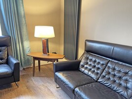 Een hotelkamer met een leren bank, een bijpassende fauteuil, een klein houten tafeltje met een lamp en een raam met gordijnen.