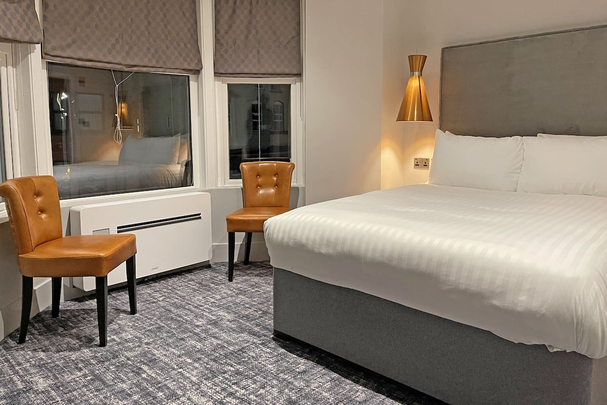 Ein Hotelzimmer mit einem Bett, zwei Stühlen, einem Fenster mit Jalousien, einer Heizung und einer Wandlampe.