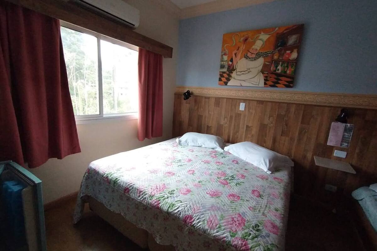 A bedroom with a wooden headboard, a bed with floral bedding, a window with red curtains, and a painting on the wall.
