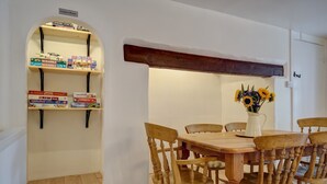 Un coin repas avec une table et des chaises en bois, une bibliothèque contenant des jeux de société et un vase rempli de tournesols.