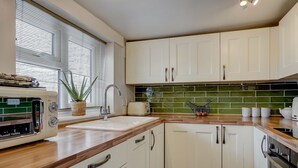 Une cuisine avec des meubles blancs, un plan de travail en bois, un grille-pain, un évier et un crédence recouverte de carreaux verts.