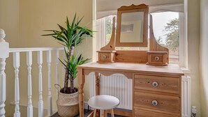 Une coiffeuse en bois avec un miroir et des tiroirs, un tabouret blanc, une plante en pot et une rambarde blanche.
