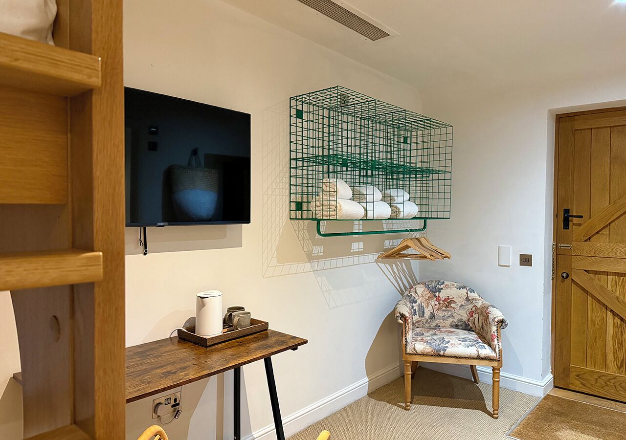 Una habitación con una silla de madera, una mesita con una tetera, un televisor montado en la pared y una cesta de alambre verde con toallas.