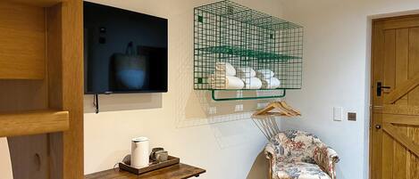 A room with a wooden chair, a small table with a kettle, a TV mounted on the wall, and a green wire basket with towels.