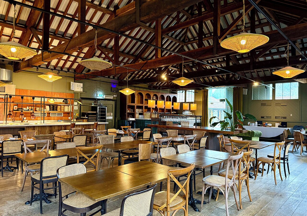 Ein geräumiges Restaurant mit hohen Decken, Holzbalken und Korbstühlen. Tische sind zum Essen gedeckt, und im Hintergrund ist eine Bar erkennbar.