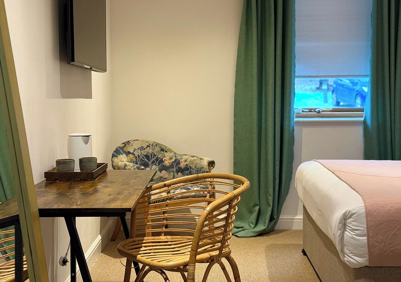 Una habitación de hotel con cama, una mesa de comedor con sillas, un televisor montado en la pared y una ventana con cortinas.