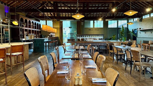 Een restaurant met houten tafels en stoelen, een bar en grote ramen.