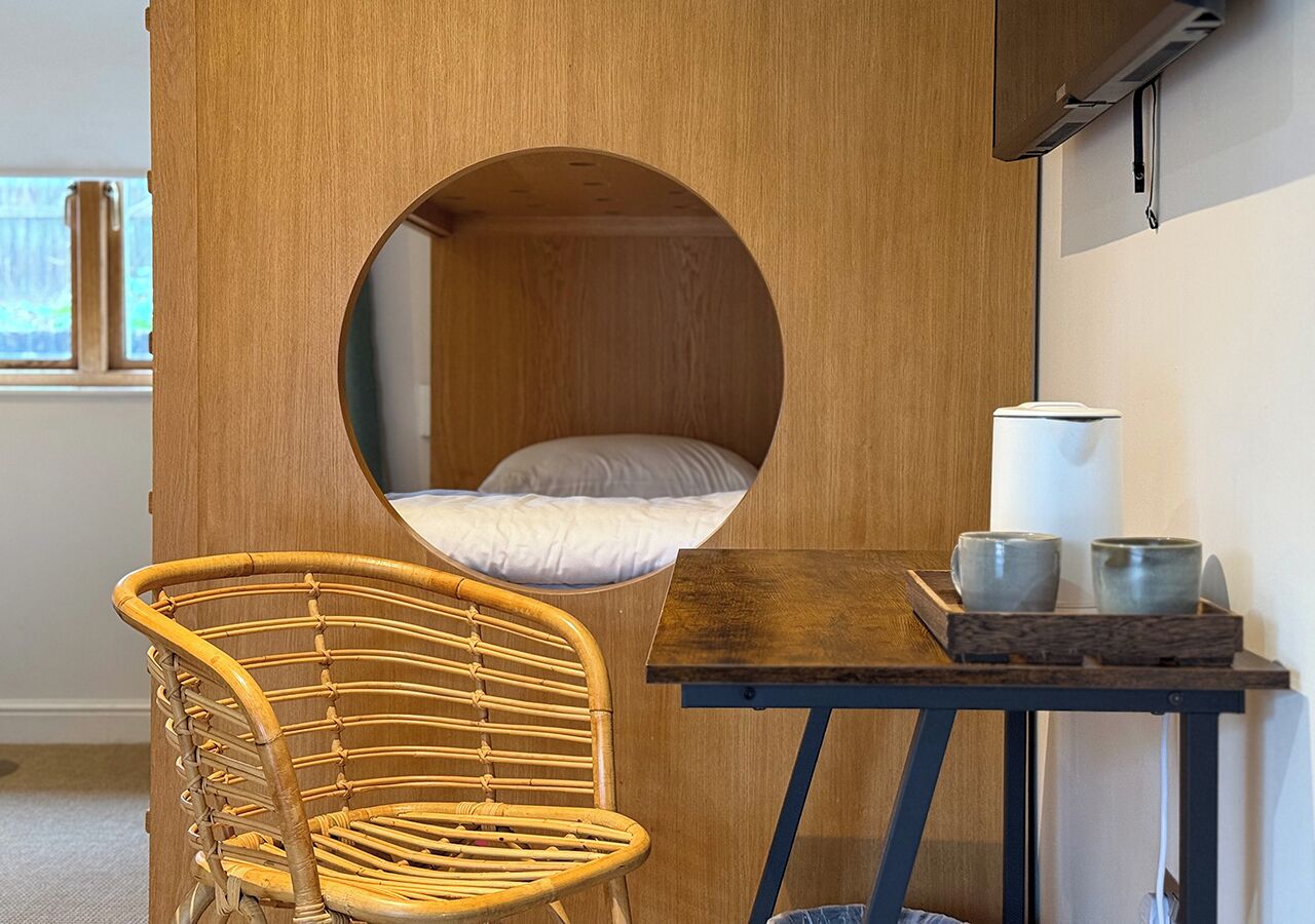 A cozy room with a circular window, a wicker chair, a small table with two cups, and a bed with white bedding.