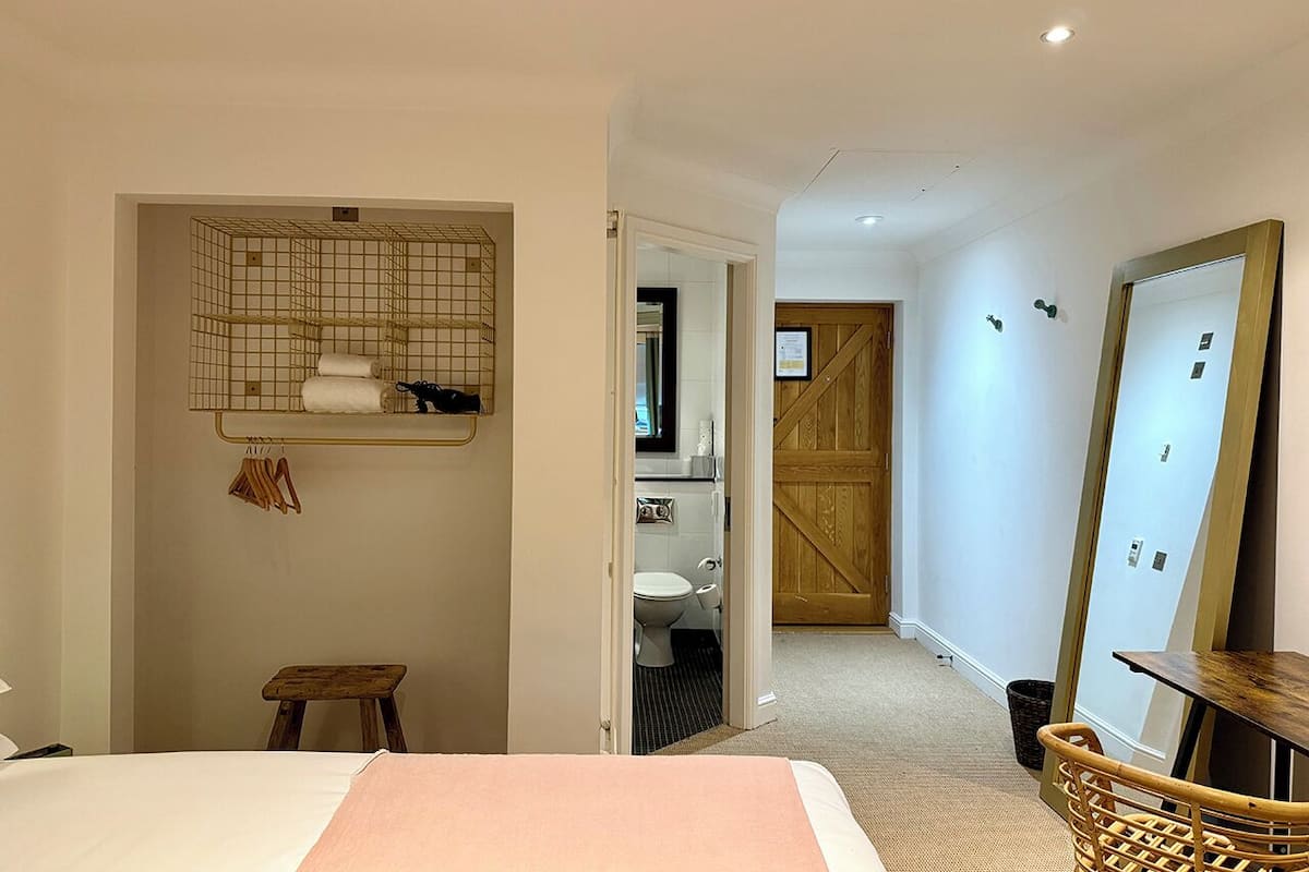 Ein Schlafzimmer mit einem Bett, einem kleinen Hocker, einem Regal mit Handtüchern, einer Holztür und einem durchgegangenen Tür erkennbaren Badezimmer.