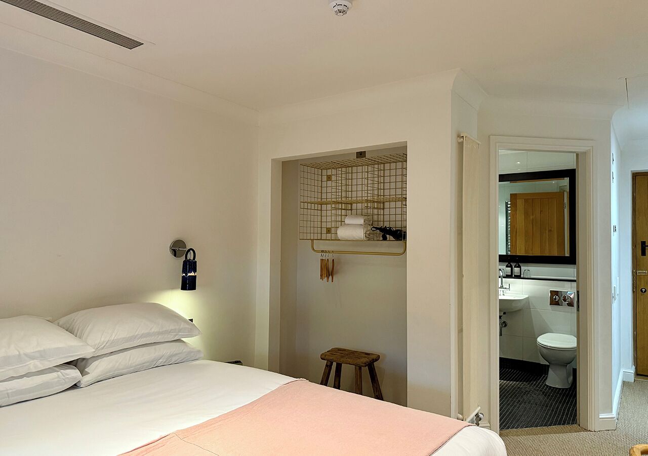 A bedroom with a bed, a nightstand, a wall-mounted shelf with toiletries, and an open door leading to a bathroom.