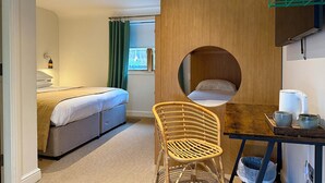 Een kamer met een bed, een rond raam, een houten scheidingswand, een rieten stoel en een tafel met waterkoker en kopjes.
