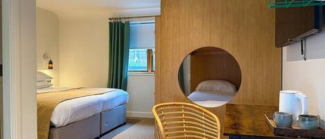 A room with a bed, a circular window, a wooden partition, a wicker chair, and a table with a kettle and cups.