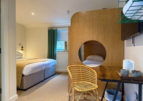 Quarto com cama, janela redonda, divisória de madeira, cadeira de vime e mesa com chaleira e xícaras.