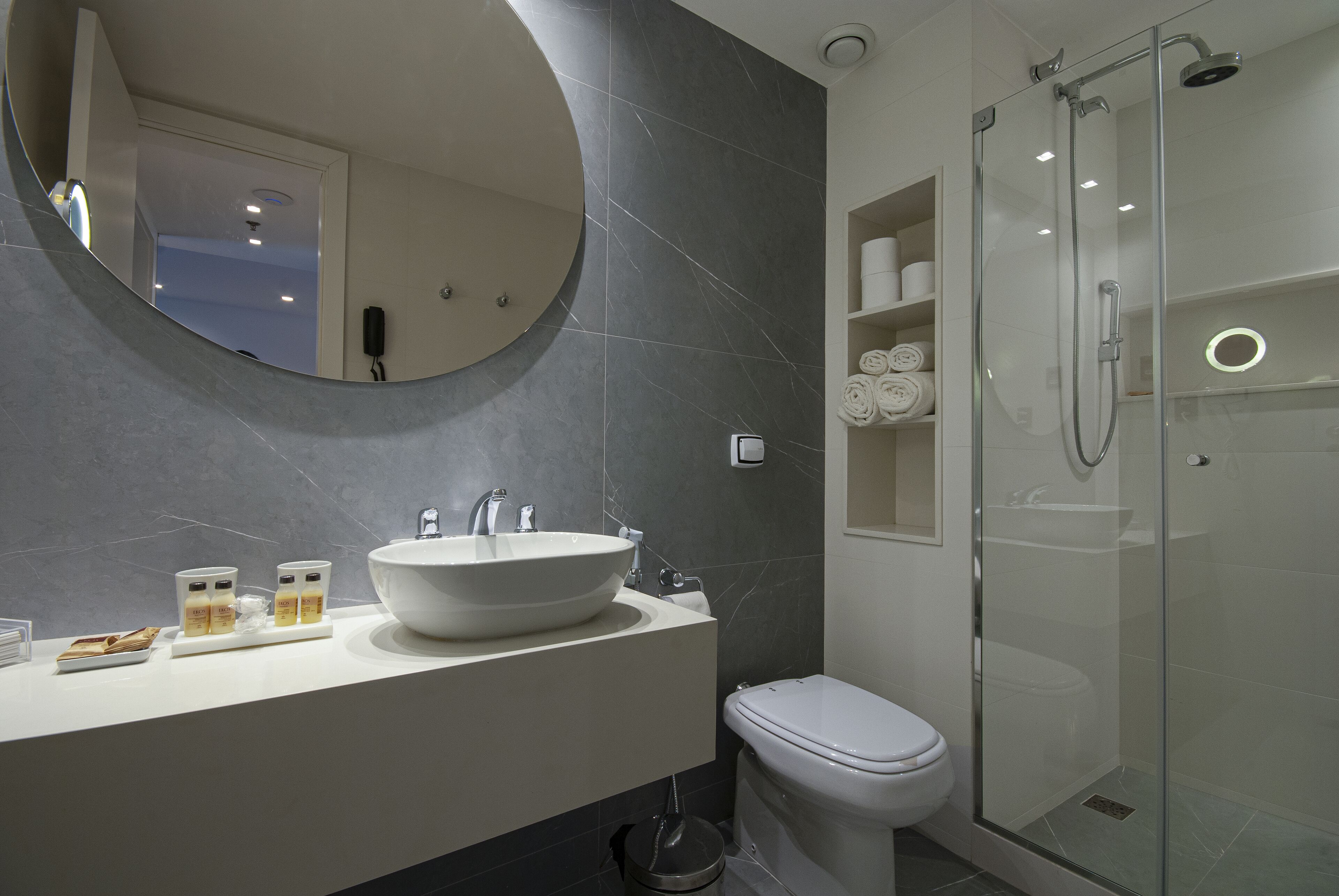 A modern bathroom with a large oval mirror, a white sink, and a glass-enclosed shower.