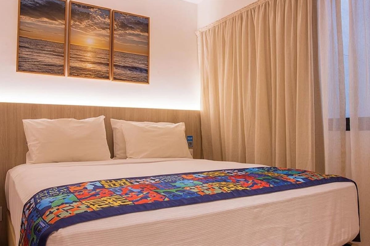 Quarto de hotel com uma cama, um edredom colorido, uma cabeceira e uma pintura do pôr do sol na parede.